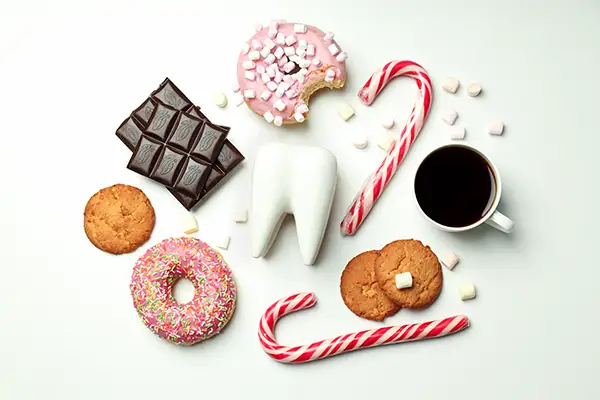 A tooth model laying on a table next to donuts, cookies, coffee, sugar cubes, chocolate, and candy canes - all examples of foods you should eat in moderation to protect the health of your teeth.