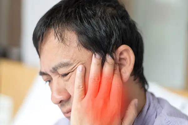 A man holding his jaw with a pained expression, red highlight on his cheek emphasizing TMJ pain.